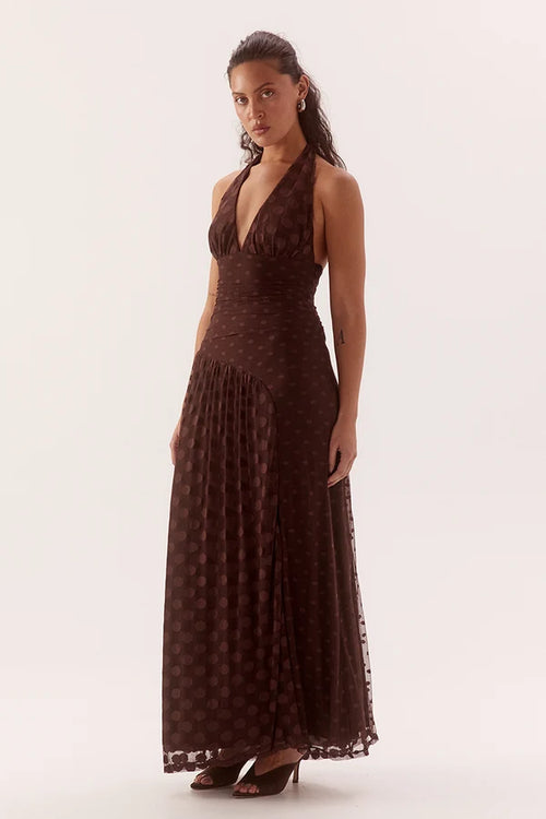 A woman modeling a dark brown halter-neck maxi dress with a subtle polka dot pattern, photographed against a plain light background.
