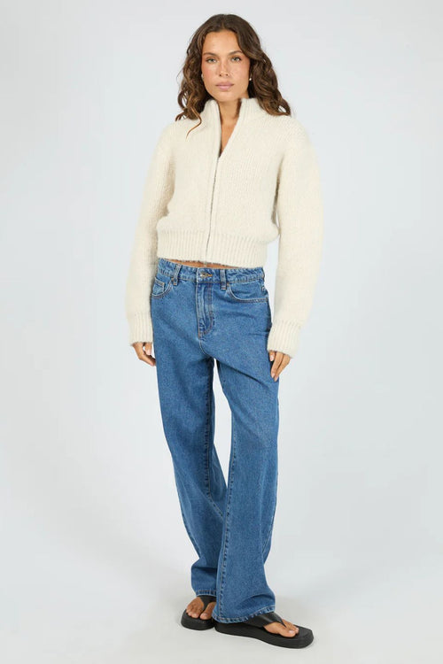 Woman wearing a cream cardigan and blue jeans on a white background