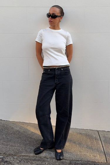 A person stands against a light gray wall wearing a fitted white short-sleeve T-shirt, loose black jeans with a studded belt, black flats, and black sunglasses.
