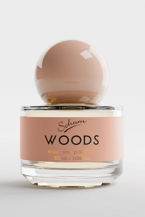 A round glass bottle of Sohum Woods eau de parfum with a glossy nude-toned spherical cap and a warm terracotta-pink label, featuring black and mustard-yellow text for an earthy, sophisticated aesthetic.