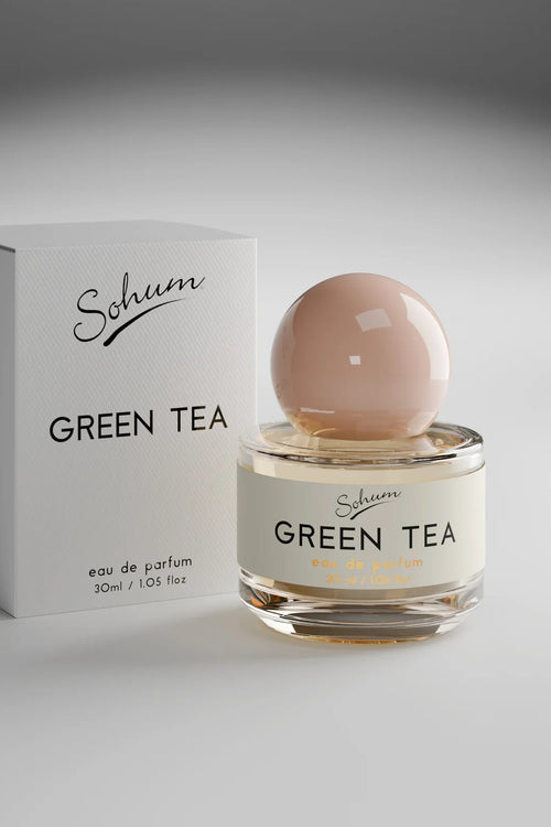 A round glass bottle of Sohum Green Tea eau de parfum with a soft blush-toned spherical lid, placed beside its minimal white textured box. The label is cream with black and amber lettering.