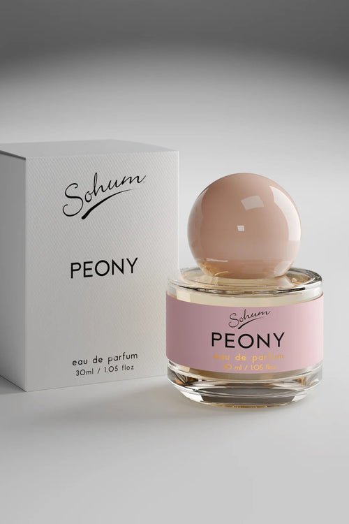 A round glass bottle of Sohum Peony eau de parfum with a blush-toned spherical cap and a soft pink label, featuring black and golden-yellow text for a fresh, feminine look.