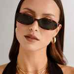 Woman wearing sunglasses and gold necklaces against a plain background