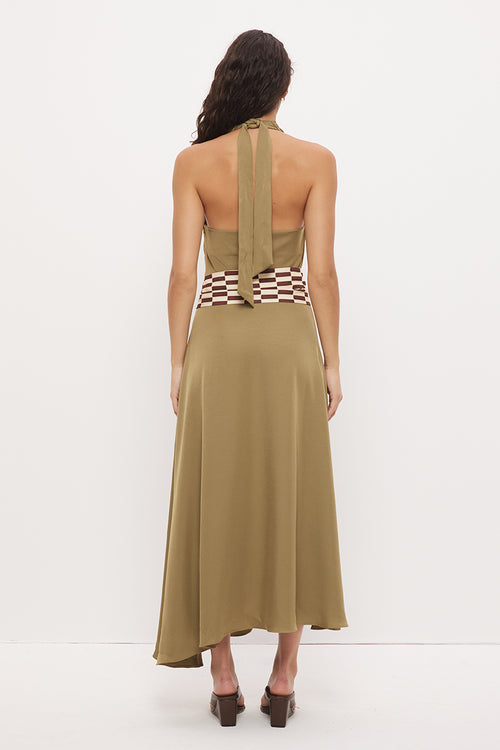 A woman viewed from the back wearing a sleeveless olive-khaki halter maxi dress with a wide checkered waistband and a long tie at the back, paired with dark brown heels against a plain white studio background.