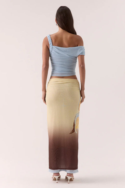 Woman wearing a light blue off-shoulder top and a long, gradient skirt on a plain background