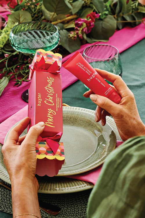 A woman at a decorated table holds a red tube of Huxter hand cream next to a red box printed with 'Merry Christmas'.