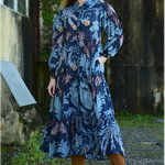 Woman wearing a blue floral dress standing in front of a rustic building.