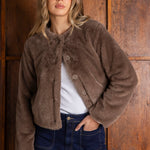 Woman wearing a brown fleece jacket and blue jeans against a wooden paneled wall.