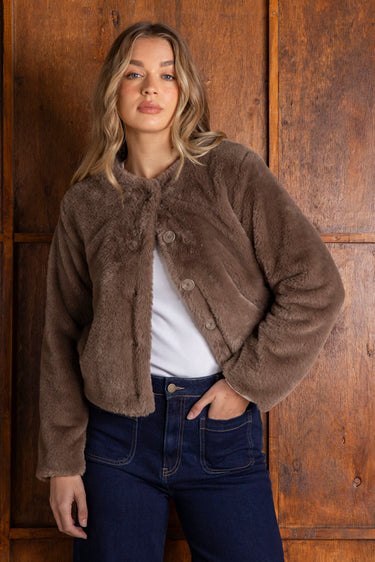 Woman wearing a brown fleece jacket and blue jeans against a wooden paneled wall.