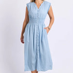A woman modeling a light blue sleeveless midi dress with a smocked waist, buttoned front, and side pockets, photographed against a plain white studio background.
