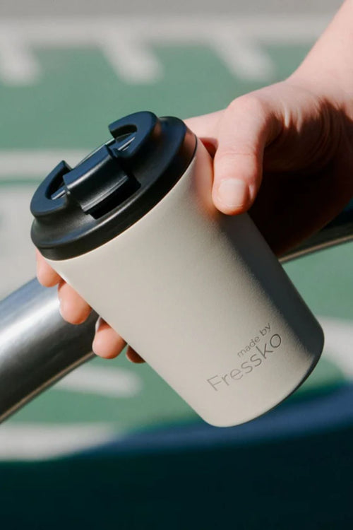 Hand holding a beige tumbler with a black lid, featuring the brand 'Fressko', against a blurred background.