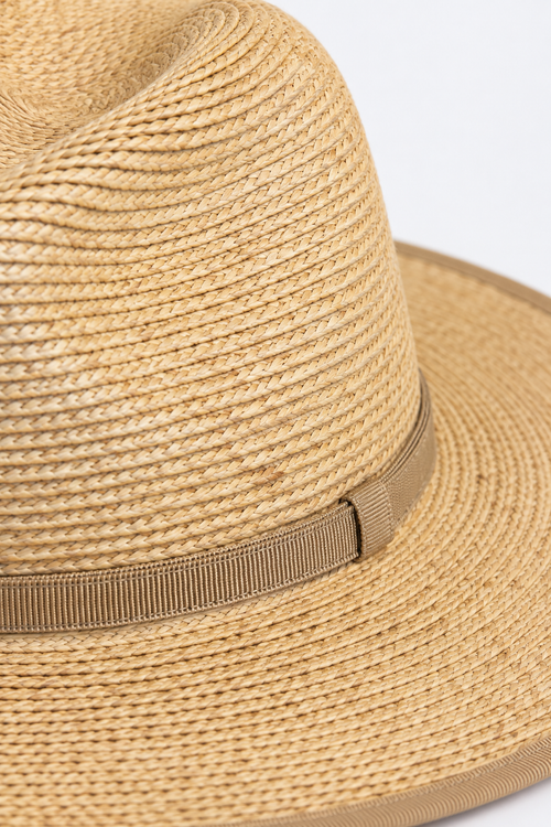 Close-up of a woven straw fedora hat in a natural tan color, featuring a ribbed tan band around the crown, set against a plain white background.