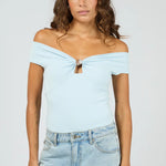A woman modeling a light blue off-the-shoulder knit top with a front twist detail and keyhole cutout, paired with light-wash jeans against a plain light gray studio background.