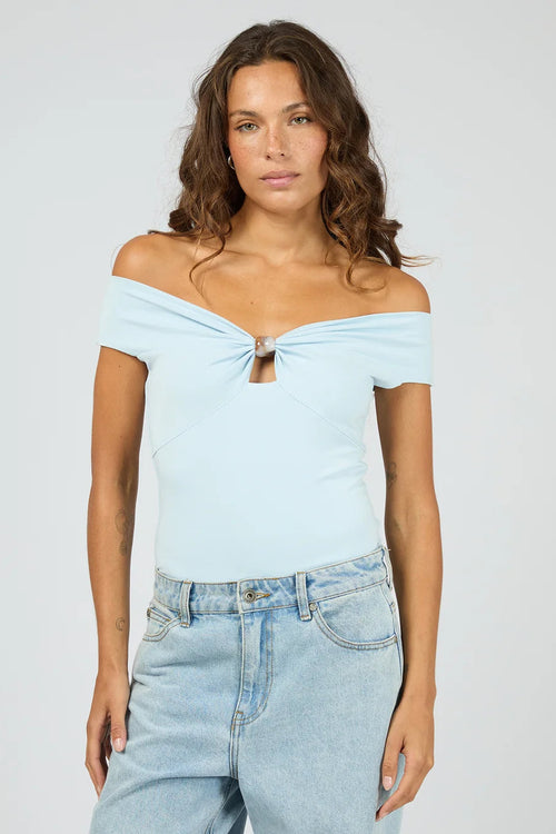 A woman modeling a light blue off-the-shoulder knit top with a front twist detail and keyhole cutout, paired with light-wash jeans against a plain light gray studio background.