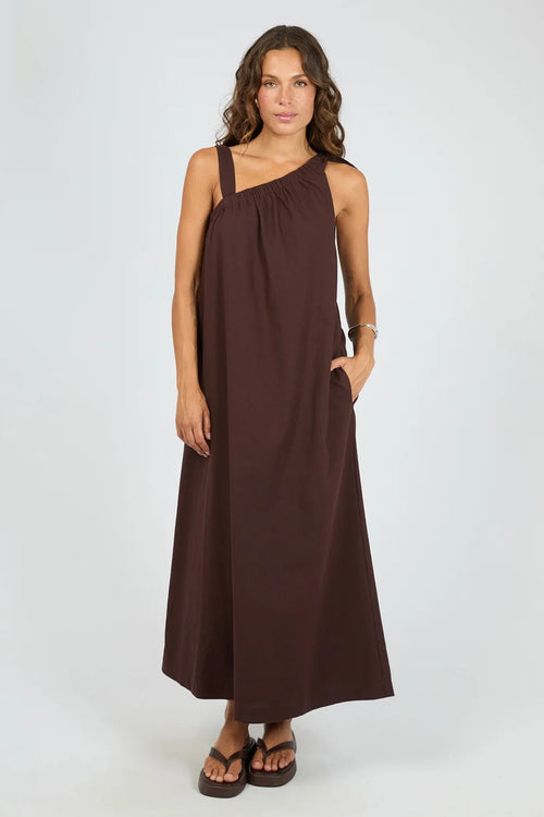 A woman modeling a dark brown one-shoulder maxi dress with a side pocket, paired with brown sandals, against a light gray studio background.