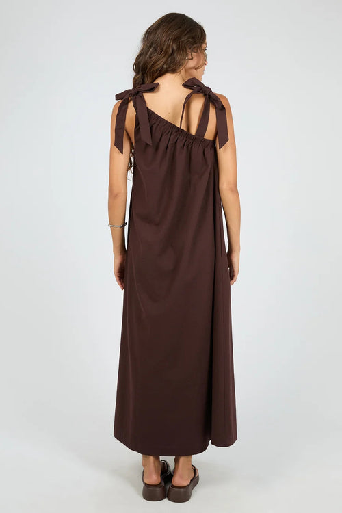 A woman viewed from the back wearing a dark brown sleeveless maxi dress with shoulder tie details and a gathered neckline, paired with dark brown flat sandals, against a light gray studio background.