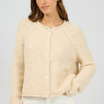 Woman wearing a beige knitted cardigan and white pants on a light gray background