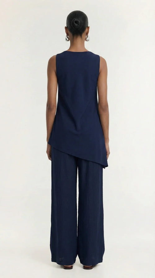 A model viewed from the back wearing a navy blue sleeveless top with an asymmetrical hem and matching wide-leg pants, set against a plain white studio background.