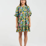A woman stands against a light gray studio background wearing a short-sleeve tiered dress with a colorful floral print, brown flat shoes, and a yellow headband.