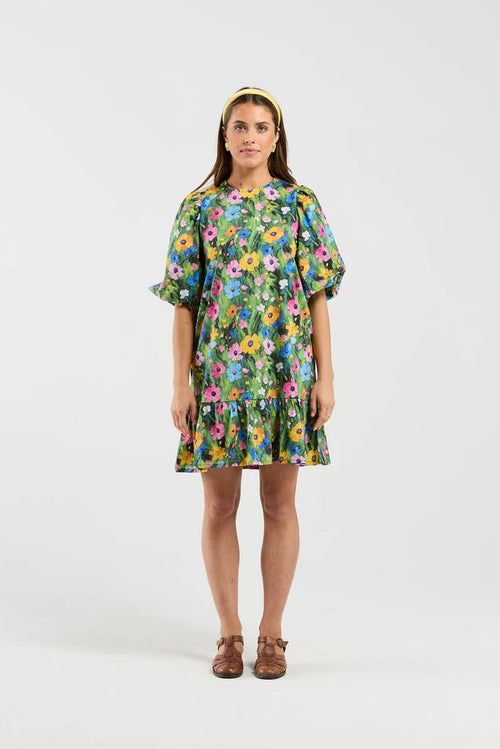 A woman stands against a light gray studio background wearing a short-sleeve tiered dress with a colorful floral print, brown flat shoes, and a yellow headband.