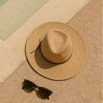 A straw sun hat and black sunglasses resting on a beige stone poolside surface next to turquoise water.