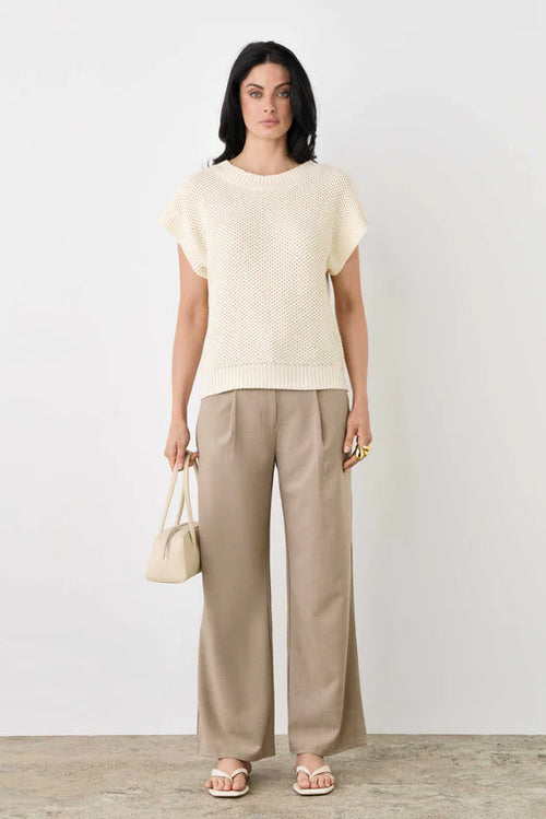 Woman wearing a cream top and beige pants holding a small bag on a plain background