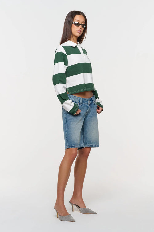A model wearing a green and white striped long-sleeve crop top, blue denim shorts, and gray pointed-toe mule heels, standing against a plain white studio background.