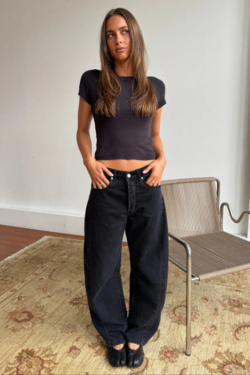 A woman stands indoors wearing a fitted dark charcoal short-sleeve crop top and very wide-leg black jeans, paired with black flats. She is posed in front of a white wall and a woven chair, with a patterned rug on the floor.