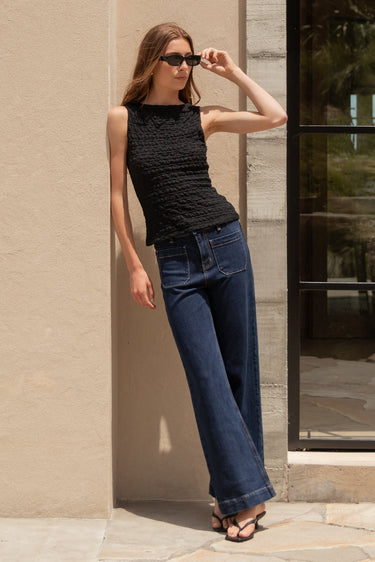 Woman in black top and blue jeans standing against a beige wall with a glass door in the background.