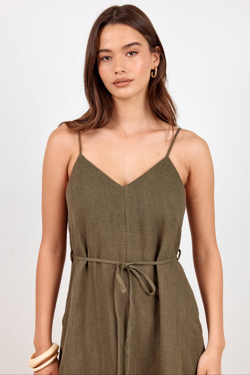 A woman modeling an olive green linen slip dress with thin spaghetti straps, a V-neckline, and a matching fabric tie belt at the waist, set against a plain light gray studio background.