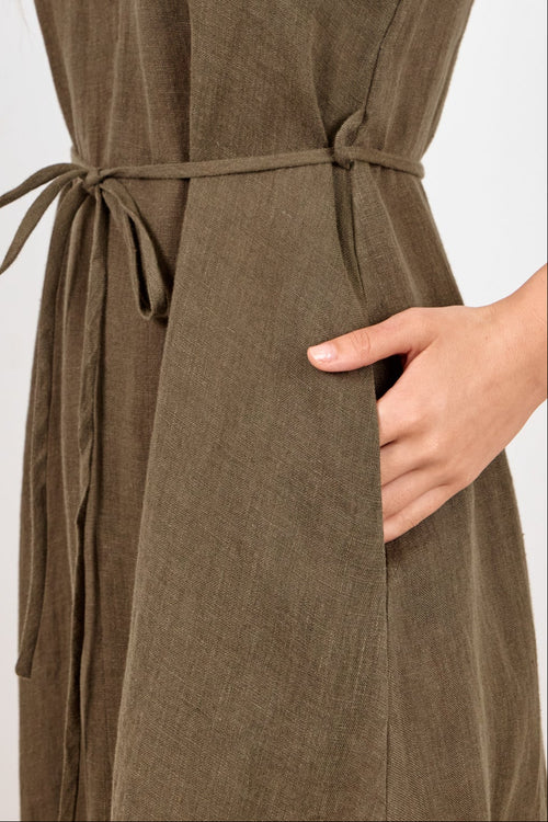 A woman wearing an olive green sleeveless midi dress with thin straps, a tie waist, and side pockets, photographed against a plain light gray studio background.