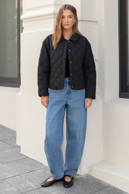 Woman wearing a black quilted jacket and blue jeans standing in front of a building.