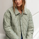 Woman wearing a green quilted jacket and blue jeans against a white wall.
