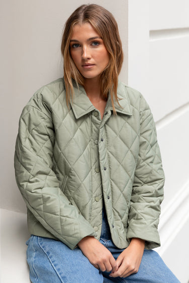 Woman wearing a green quilted jacket and blue jeans against a white wall.