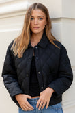 Woman wearing a black quilted jacket and blue jeans against a light-colored wall.