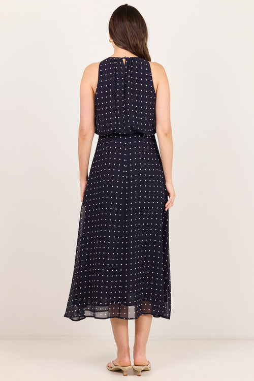 A woman viewed from the back wearing a sleeveless navy midi dress with a small white polka dot pattern, standing against a plain off-white studio background.