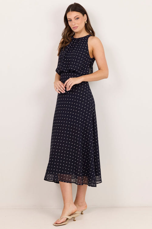 A woman modeling a sleeveless navy midi dress with a small white polka dot pattern, standing against a plain off-white studio background.