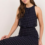 A woman wearing a sleeveless navy maxi dress with a white polka dot pattern, sitting on a white stool against a plain off-white background.