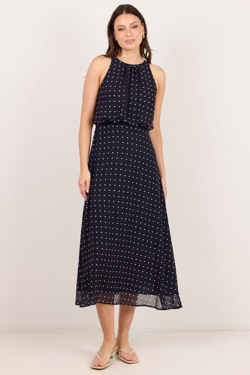 A woman modeling a sleeveless navy midi dress with a white polka dot pattern, standing against a plain off-white studio background.
