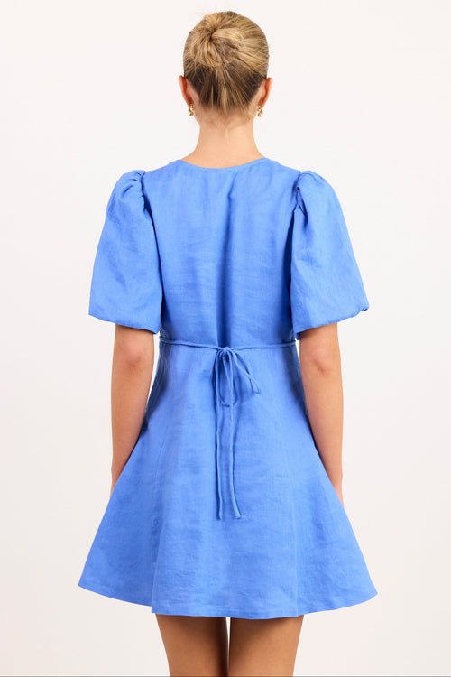 A woman viewed from the back wearing a short-sleeve blue dress with puffed sleeves and a tie at the waist, photographed against a plain light background.