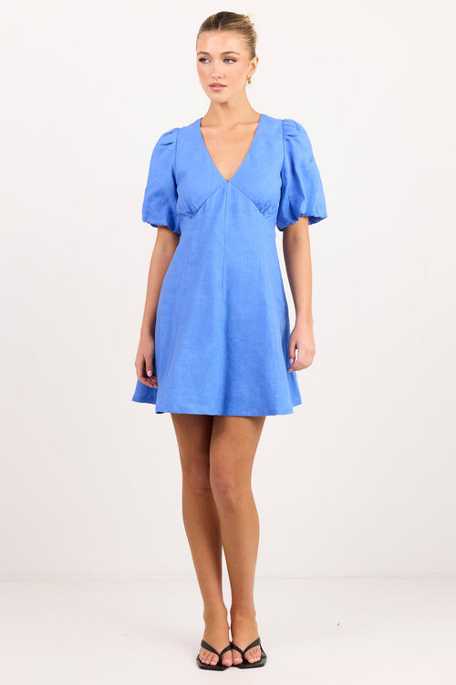 A woman modeling a short blue wrap-style dress with puff sleeves and a V-neckline, standing against a plain white studio background.