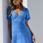 A woman wearing a bright blue linen dress with puff sleeves and a V-neckline, posing in a white architectural setting.