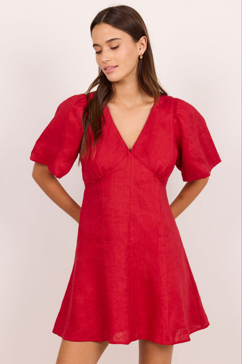 A woman modeling a red short puff-sleeve dress with a V-neckline, photographed against a plain light beige studio background.