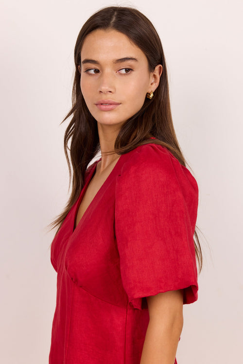 A woman with long dark brown hair is shown in a side profile wearing a red short-sleeve wrap dress against a plain light beige background.