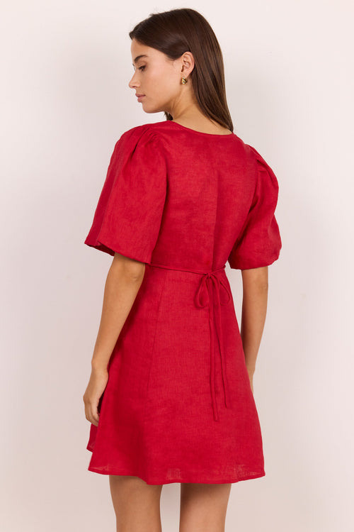 A woman modeling a red short puff-sleeve dress from the back, featuring a tie waist and textured fabric, set against a plain light background.