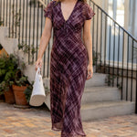 A woman wearing a dark purple and brown plaid midi dress with flutter sleeves walks down a brick courtyard path next to a building with black metal stairs.