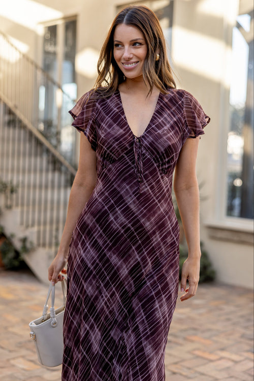 A woman wearing a dark purple and black plaid midi dress with flutter sleeves and a V-neckline, holding a small white bucket bag in an outdoor patio setting.