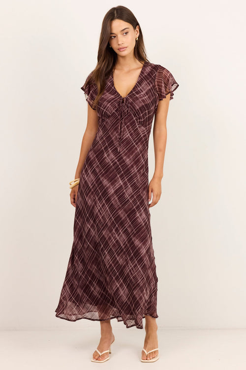 A woman modeling a dark plum purple and brown plaid maxi dress with flutter sleeves and a V-neckline, standing against a plain off-white studio background.