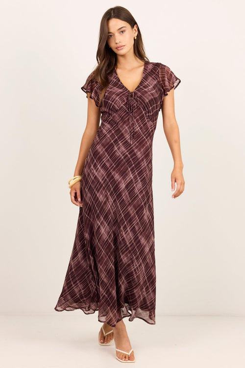 A woman modeling a long, dark plum maxi dress with a subtle plaid pattern, flutter sleeves, and a V-neckline, standing against a plain off-white studio background.