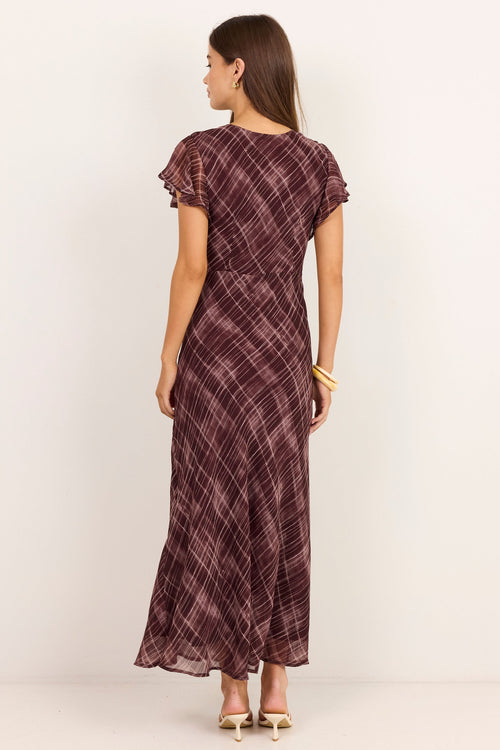 A woman stands with her back to the camera wearing a dark plum plaid maxi dress with flutter sleeves and gold heels, set against a plain off-white studio background.